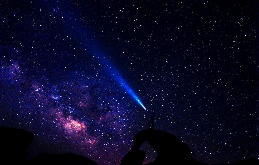 Milky way and starry sky, black silhouette of a person standing on a boulder flashing a beam of light into the dark night sky
