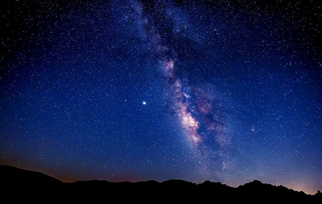 Night photo of Milky Way and starry sky with a thin band of black hilly landscape on the bottom