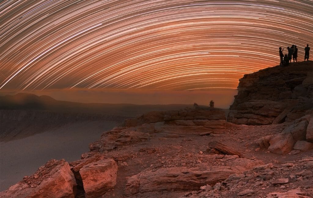 Orange sky with half round star trails, red rocks in the foreground and to the right is a boulder with the small outlines of people standing on it.