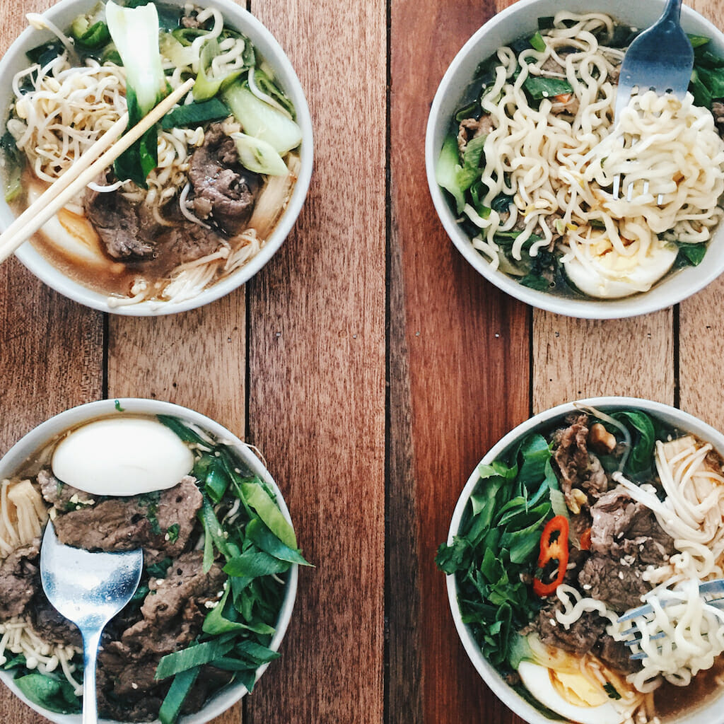 Four separate bowls of different ramen in the four corners of a brown wood table with chopsticks and spoons