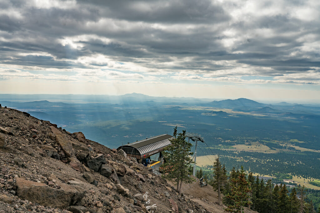 steep gravel slope in the foreground, metal roof of the gondola station in the center and vast view of the valley and dramatic sky in the background