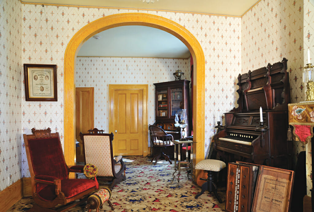 A parlor with cream and yellow wallpaper decorated with warm colored wood furniture 