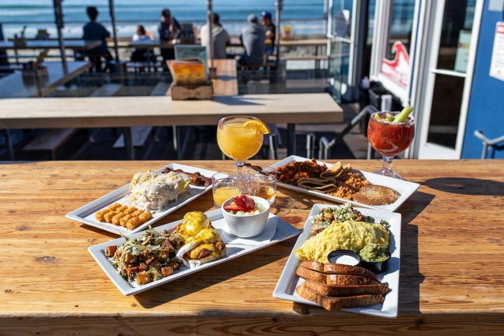 Several plates filled with breakfast foods near glasses of mimosas are displayed on a table with more chairs and the beach in the background