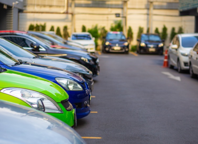 A row of parked cars in a parking lot, showcasing various colors such as green, silver, black, and blue. The outdoor lot meets minimum parking requirements and features a building and trees in the background.