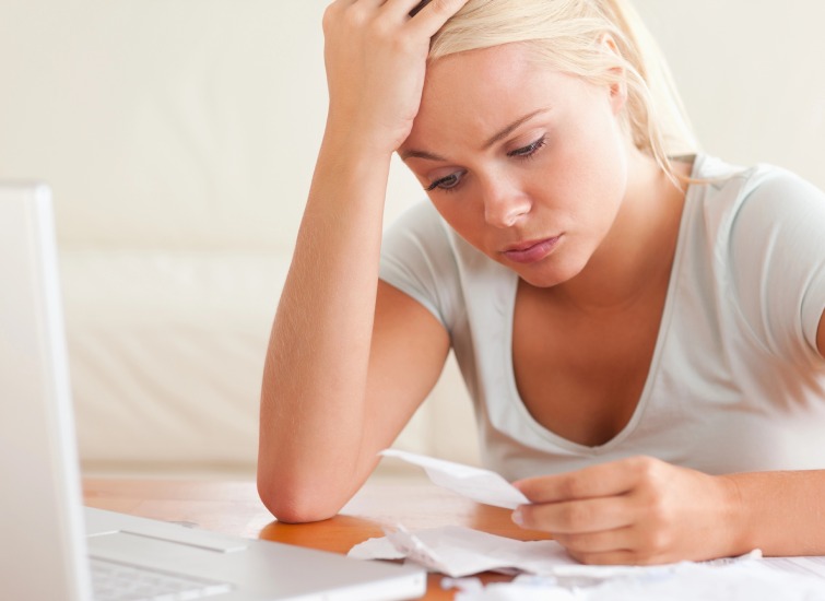 A woman with blonde hair looks stressed while holding a piece of paper. She is sitting at a table with crumpled papers and a laptop in front of her.