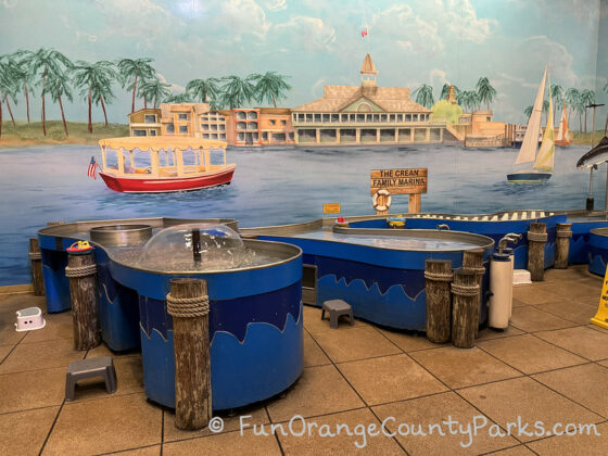 a mural depicting balboa peninsula pavilion building with an electric boat and sailboat in the harbor behind blue pools and fountains for children to play with floating objects at child level