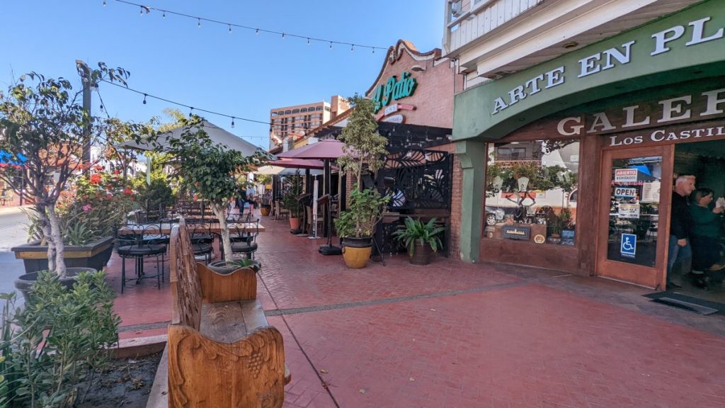 Brick sidewalk lined by shops and outdoor seating under cafe string lights and trees on sunny day. Avenida Lopez Mateos, Ensenada Sights.