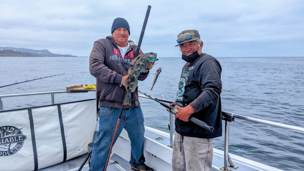 Two men on fishing boat holding fishing poles and one large fish, in background is calm water and distant mountains on gloomy day. Deep sea fishing, What to do in Ensenada.