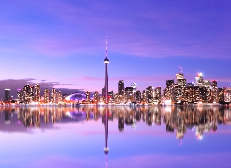 Toronto city skyline at dusk, with the CN Tower and modern buildings reflecting in calm water under a purple and blue sky.