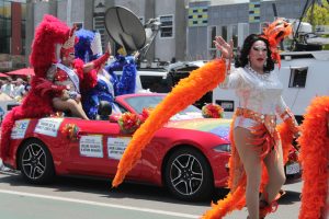 A drag queen in sparkling silver with long orange feathers waves ahead of a red converticle with matching drag queens in red and blue.