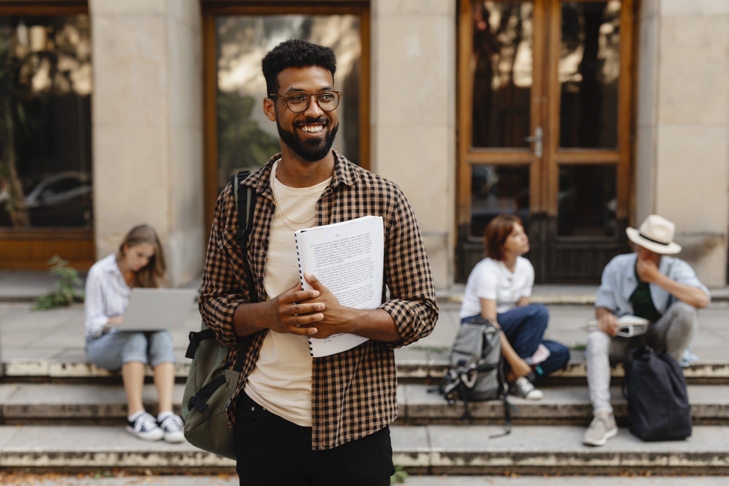 Happy young university student with book standing outdoors in front of campus.