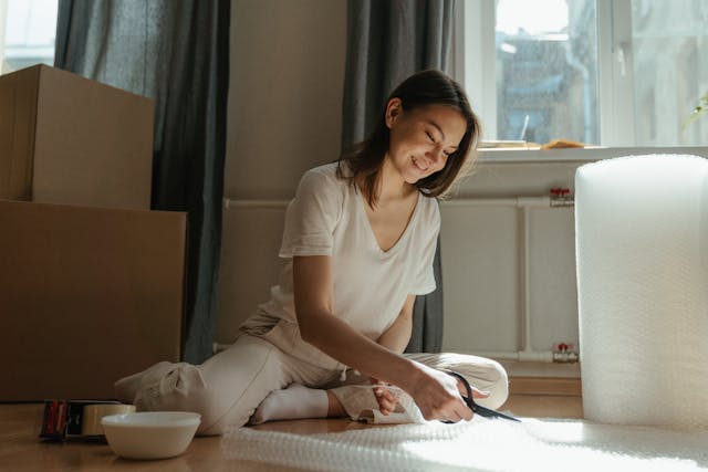 Woman sitting on the floor, surrounded by wooden boxes while cutting bubble wrap.