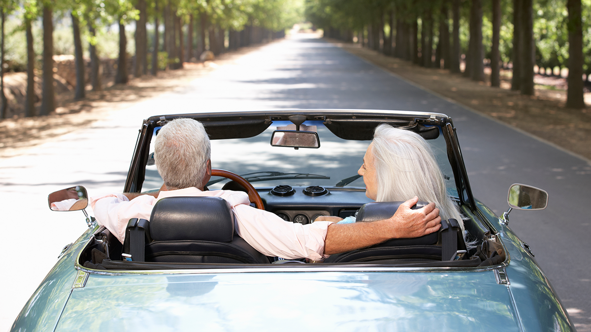 Retired couple in a sports car driving down a tree-lined street