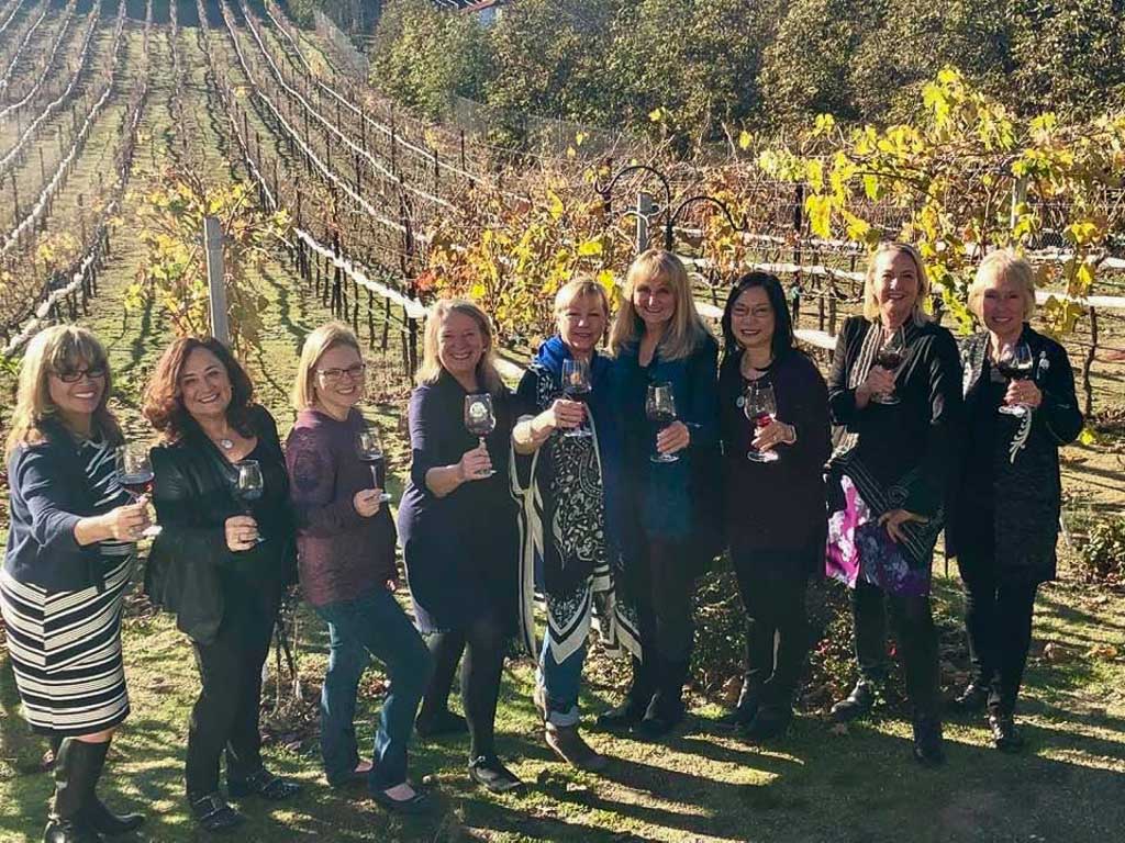 9 female wine makers standing in front of vineyard with wine