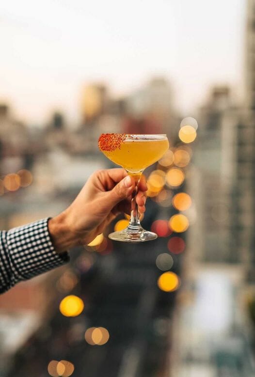 Male hand holding an orange colored cocktail in front of an out of focus city scape in the background