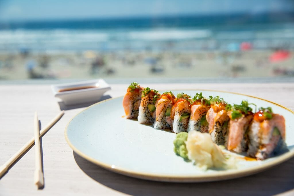 white plate with sushi roll and chopsticks on a table in the foreground. Background: out of focus, beach, people and ocean