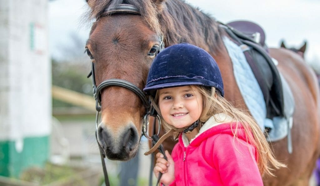 Young girl with riding helmet and pink jacket leading a horse, smiling into the camera