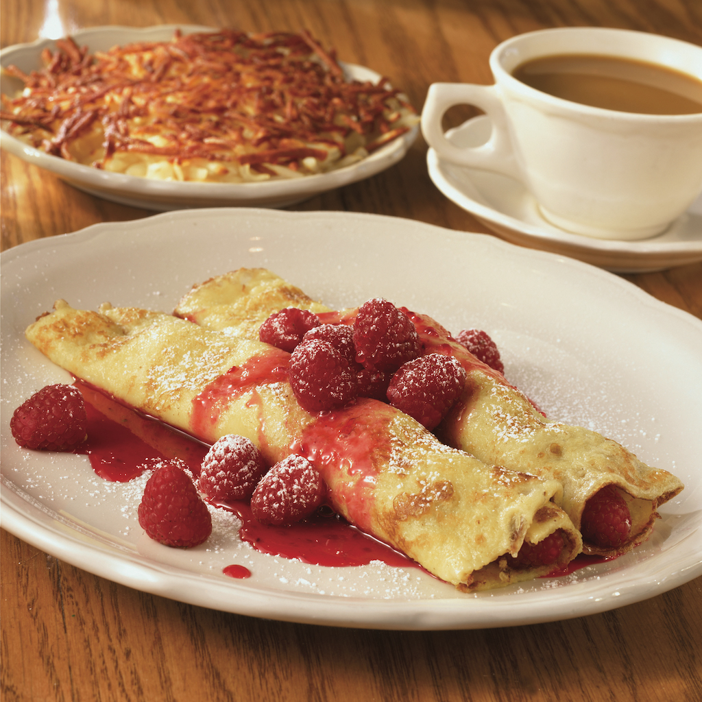 Two rolled crepes laying side by side with a raspberry topping in front of another dish and a cup of coffee
