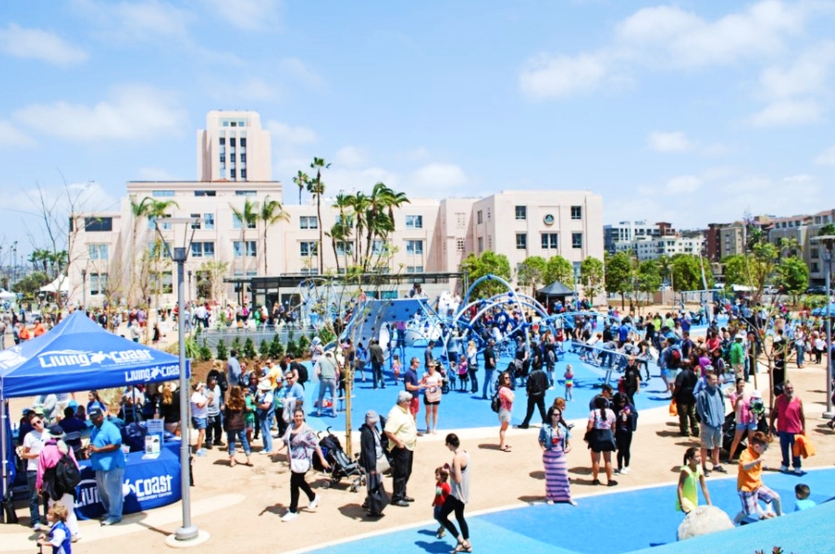 A building is the backdrop for a park with grassy spaces and many people milling about.