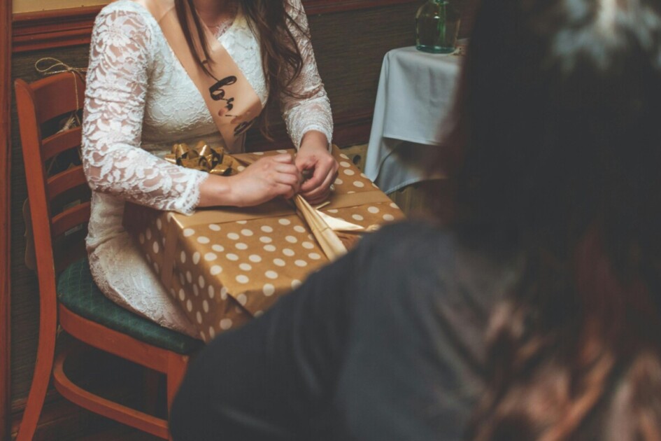 the bride-to-be wears a pink sash and opens a present in a chair at a home bridal shower