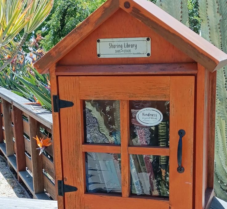 A small wooden sharing library with a glass door, filled with books, set against a garden backdrop with a cactus and bird of paradise flower.