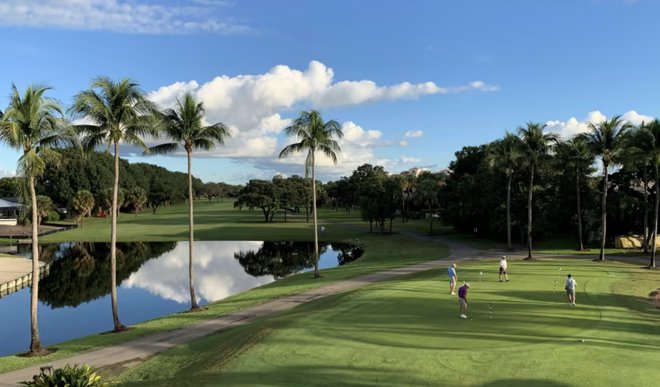 Golfers playing on a green at a Boca Raton golf course with palm trees, a reflective lake, and a bright blue sky in the background.