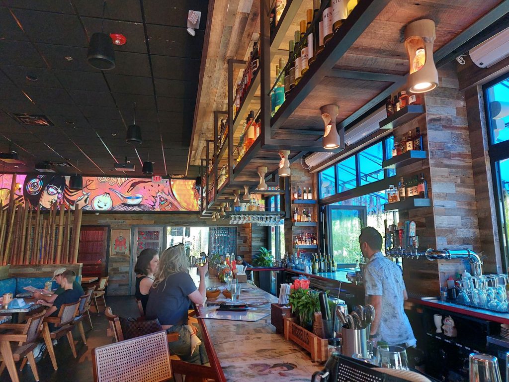 Modern restaurant and bar interior in Mizner Park with guests seated at the bar, hanging shelves filled with bottles, warm lighting, and large windows letting in natural light.