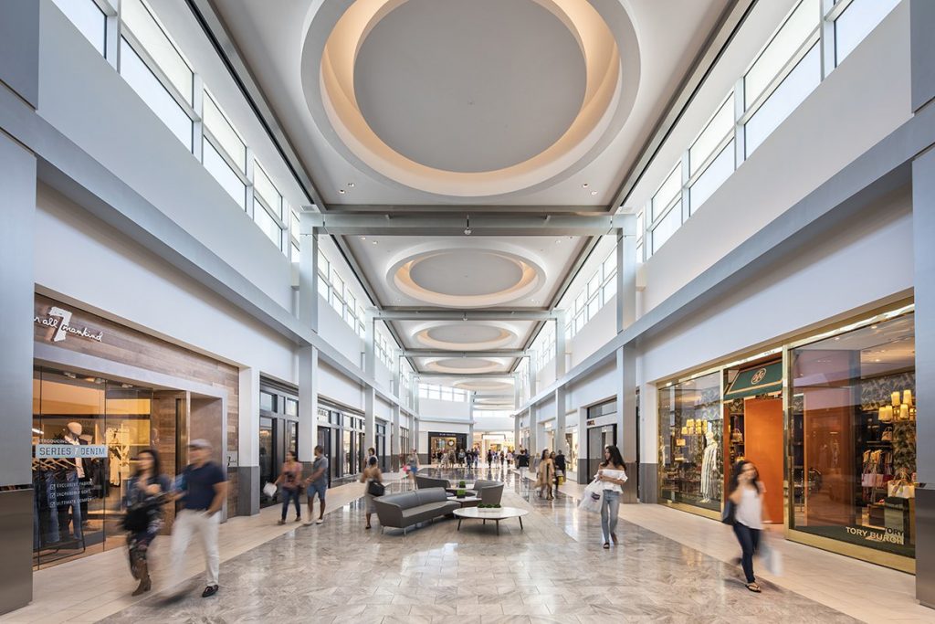 Shoppers walk through a bright, modern indoor mall with high ceilings, large circular light fixtures, and upscale retail stores on both sides.