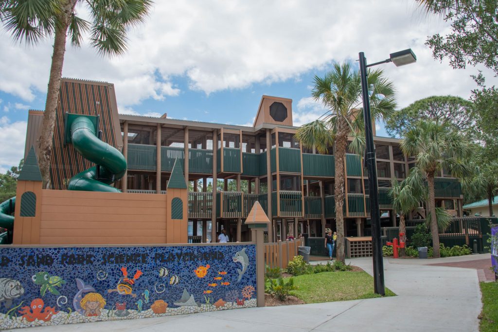 Science-themed playground structure at Sugar Sand Park in Boca Raton, Florida, featuring a large green spiral slide, castle-style towers, and colorful mosaic wall art with ocean animals.
